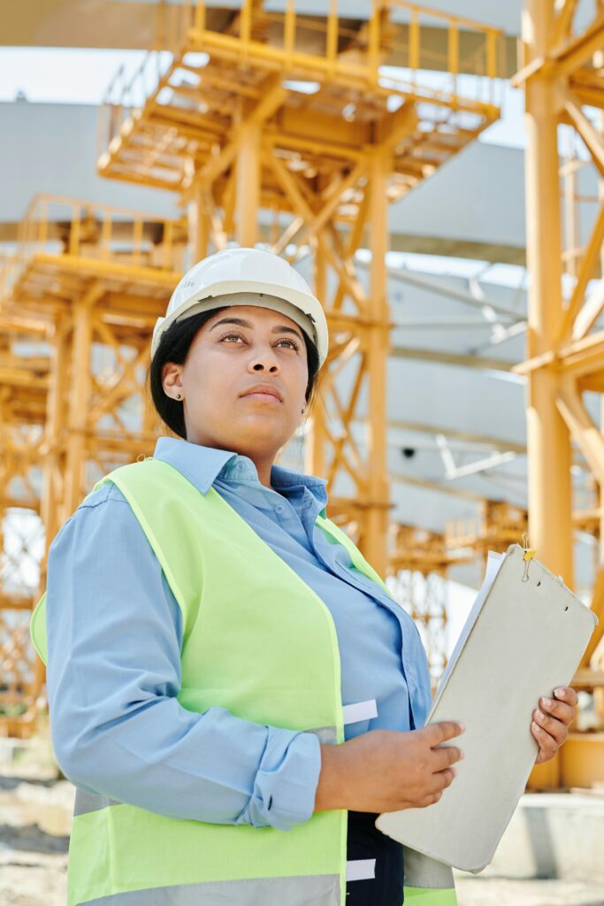 Home Confident female engineer in safety gear at a busy construction site, holding a clipboard.